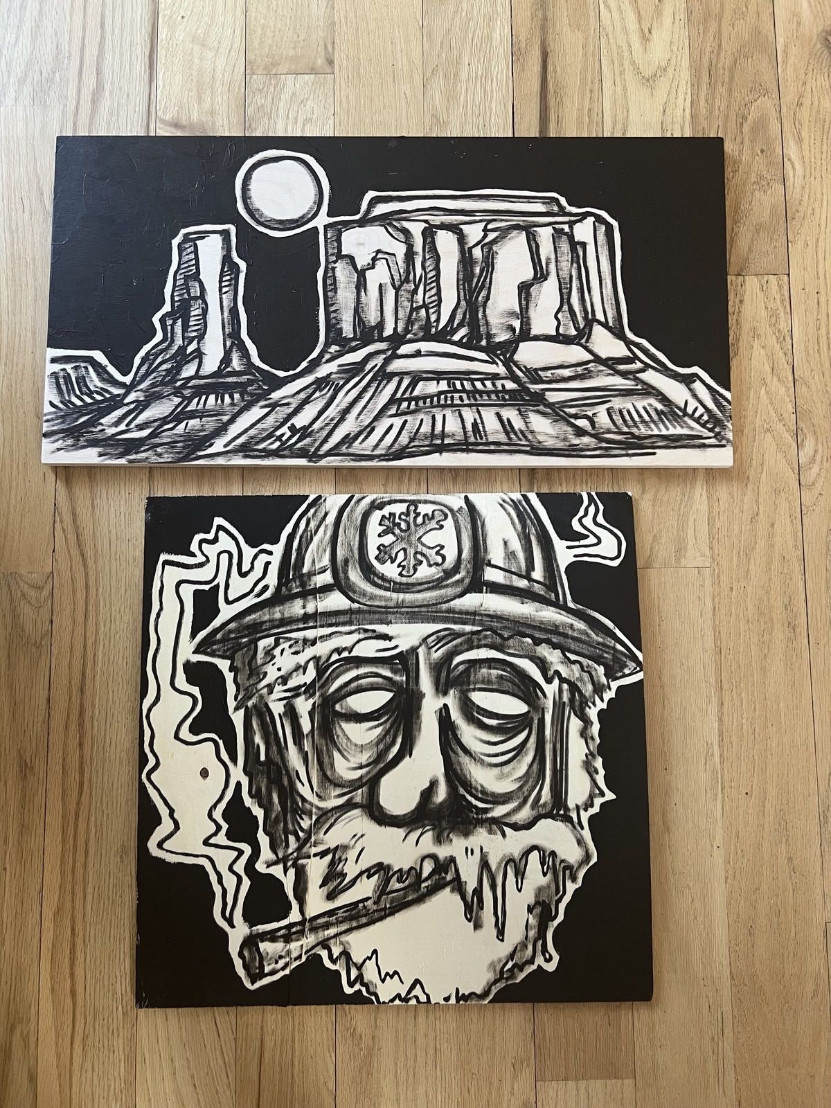 Two black-and-white acrylic panels on wood: a moonlit desert butte and a helmeted face with a cigarette